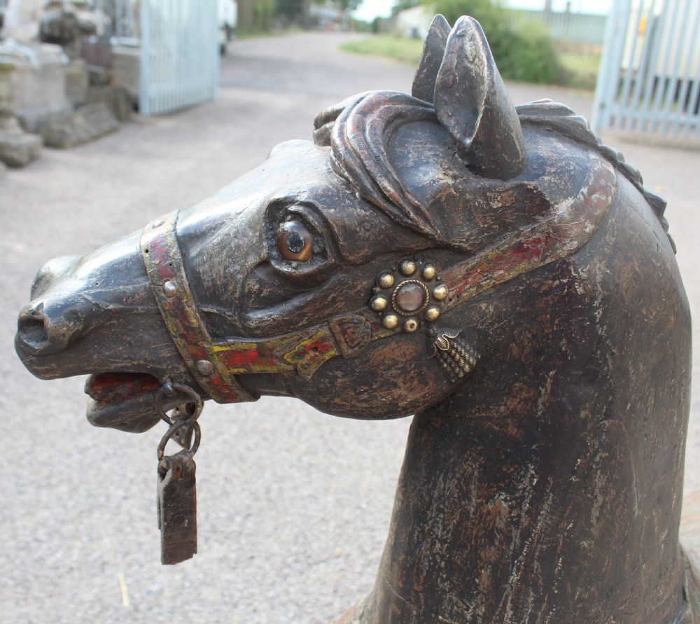 A hand-carved wooden carousel horse featuring weathered dark paint, decorative red and green saddle details, and a raised front leg pose. View XI