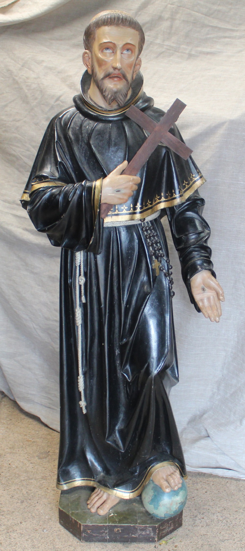 A polychrome carved wooden figure of a tonsured saint in a black hooded habit with gold trim, holding a crucifix with one foot resting on a globe.