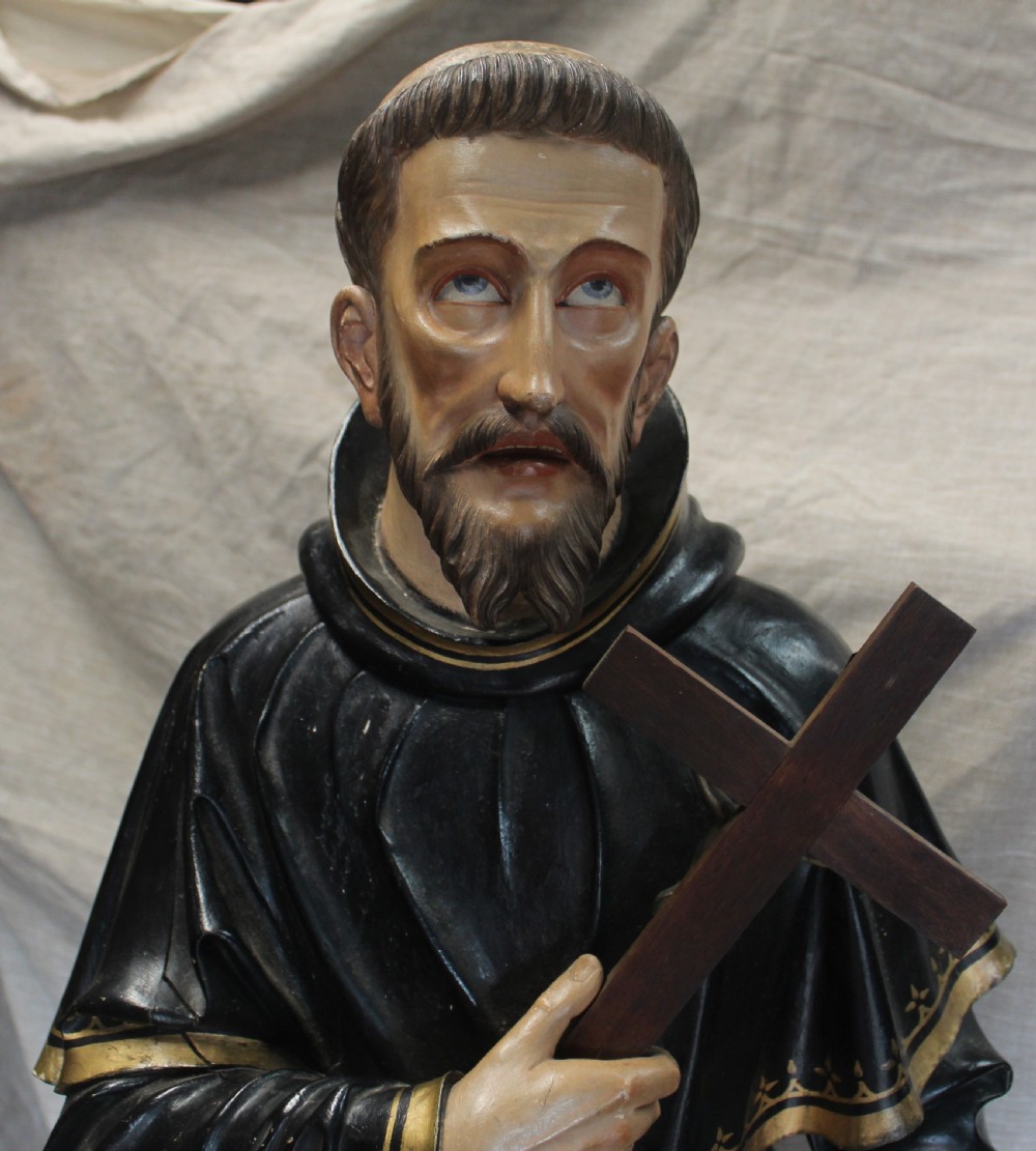 A polychrome carved wooden figure of a tonsured saint in a black hooded habit with gold trim, holding a crucifix with one foot resting on a globe. View II