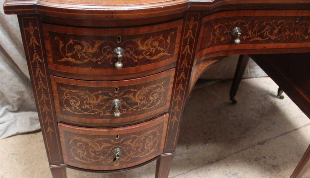 An Edwardian mahogany writing desk featuring intricate floral marquetry, a red gilt-tooled leather writing surface, a raised superstructure with drawers, and tapered legs on brass castors. View IV