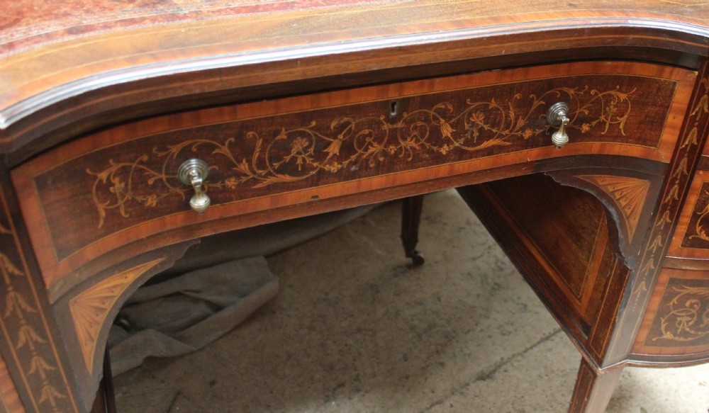 An Edwardian mahogany writing desk featuring intricate floral marquetry, a red gilt-tooled leather writing surface, a raised superstructure with drawers, and tapered legs on brass castors. View V