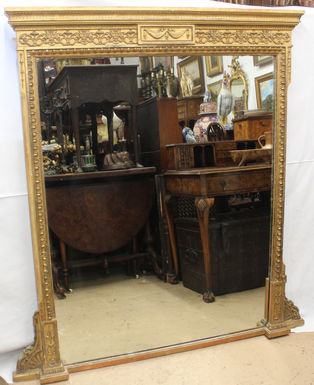 A large rectangular gilded overmantel mirror featuring a moulded frame with floral swags, a guilloche frieze, and decorative plinth bases.