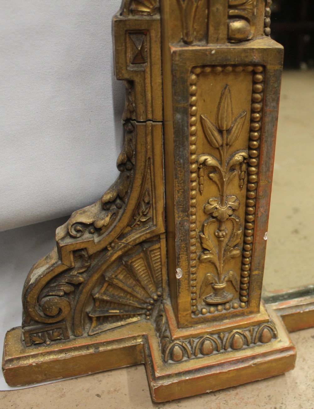 A large rectangular gilded overmantel mirror featuring a moulded frame with floral swags, a guilloche frieze, and decorative plinth bases. View V