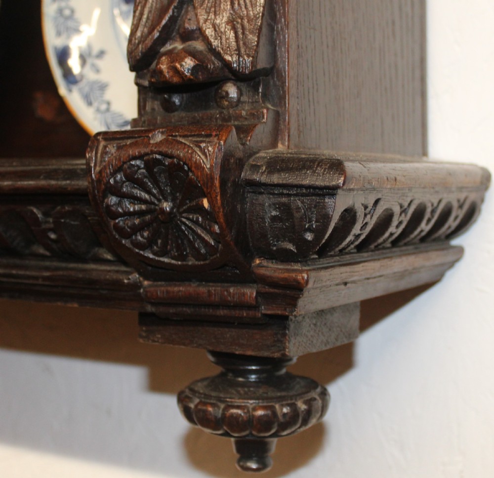 A dark carved oak wall-mounted plate rack featuring three shelves, flanked by carved caryatid figures and topped with a moulded ornate cornice. View V