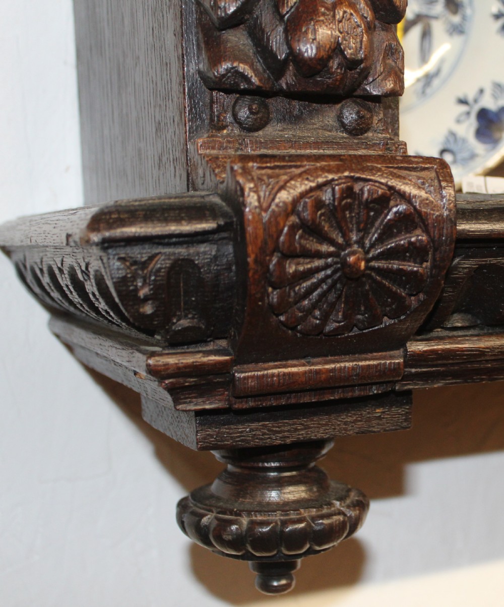 A dark carved oak wall-mounted plate rack featuring three shelves, flanked by carved caryatid figures and topped with a moulded ornate cornice. View VI