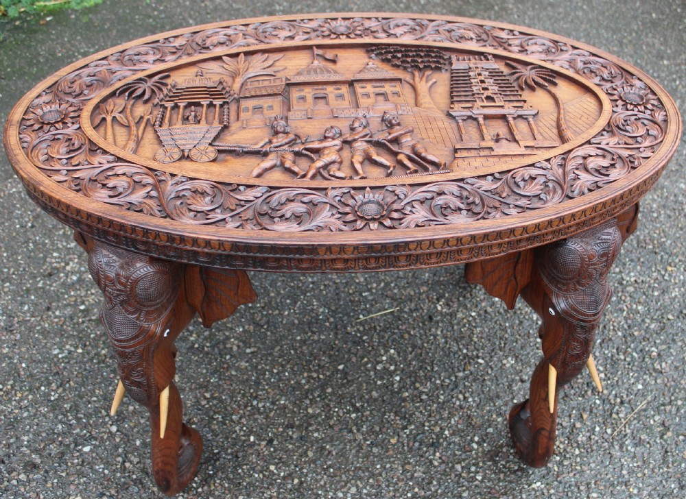An oval teak low table featuring deep relief carvings of figures and architecture on top with legs carved in the shape of elephant heads.