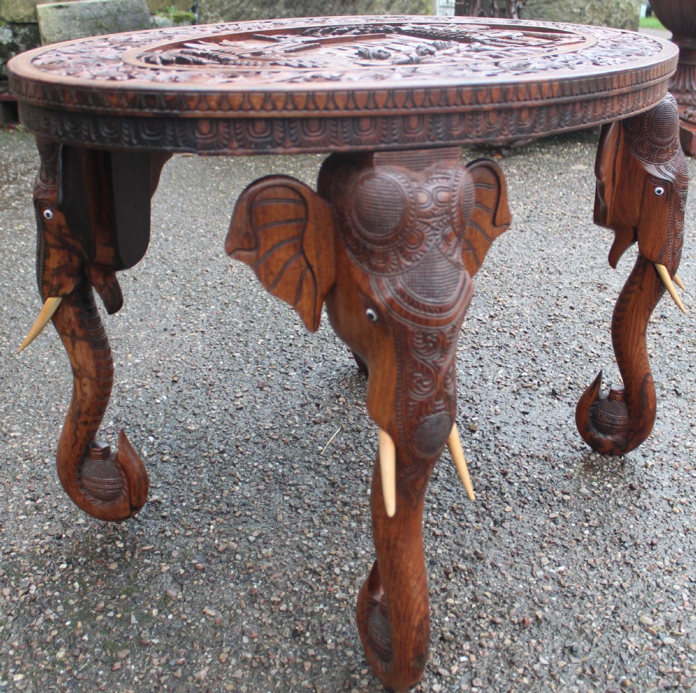 An oval teak low table featuring deep relief carvings of figures and architecture on top with legs carved in the shape of elephant heads. View II