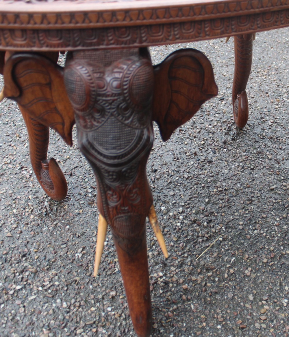 An oval teak low table featuring deep relief carvings of figures and architecture on top with legs carved in the shape of elephant heads. View III