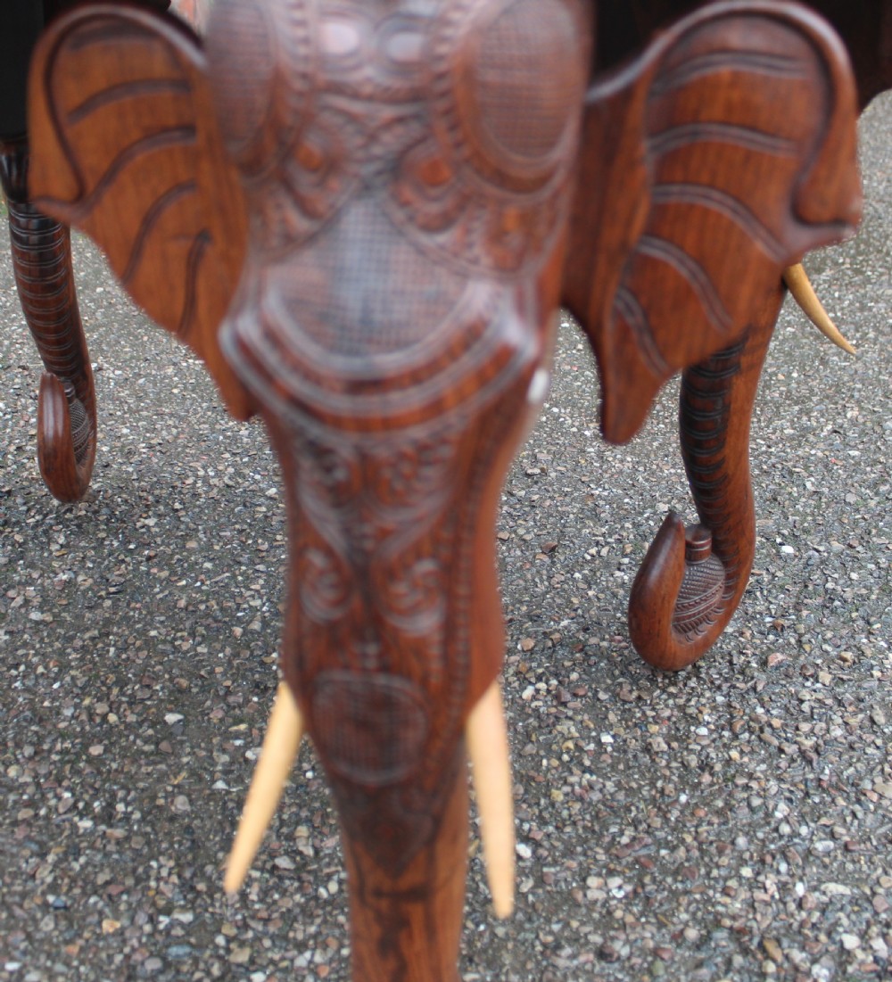 An oval teak low table featuring deep relief carvings of figures and architecture on top with legs carved in the shape of elephant heads. View IV