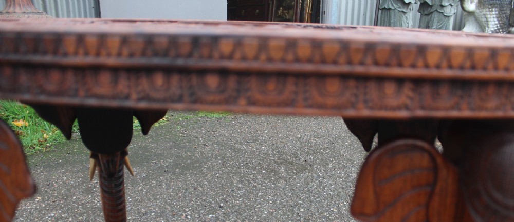 An oval teak low table featuring deep relief carvings of figures and architecture on top with legs carved in the shape of elephant heads. View V