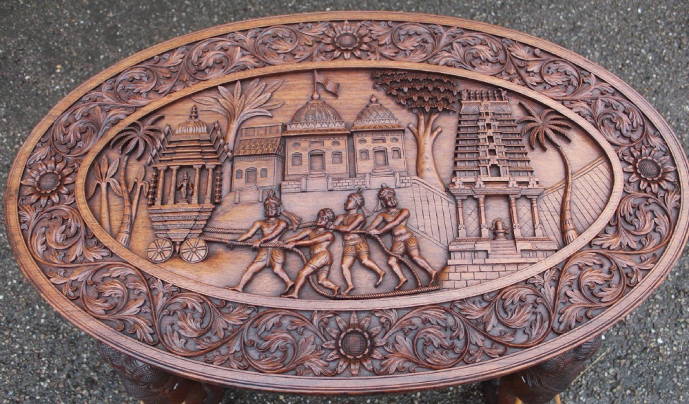 An oval teak low table featuring deep relief carvings of figures and architecture on top with legs carved in the shape of elephant heads. View VI