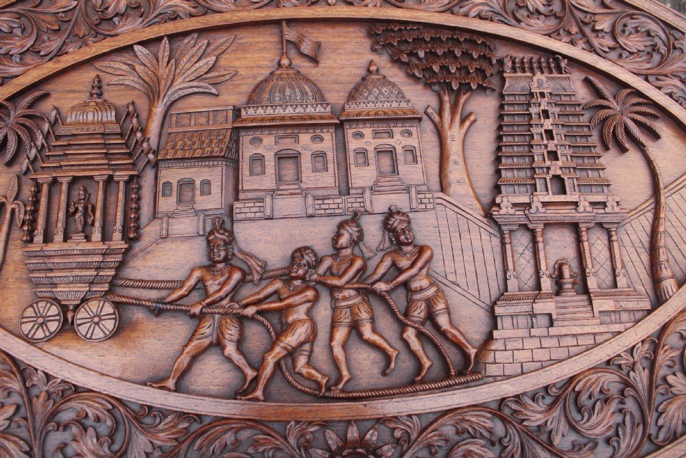 An oval teak low table featuring deep relief carvings of figures and architecture on top with legs carved in the shape of elephant heads. View VII