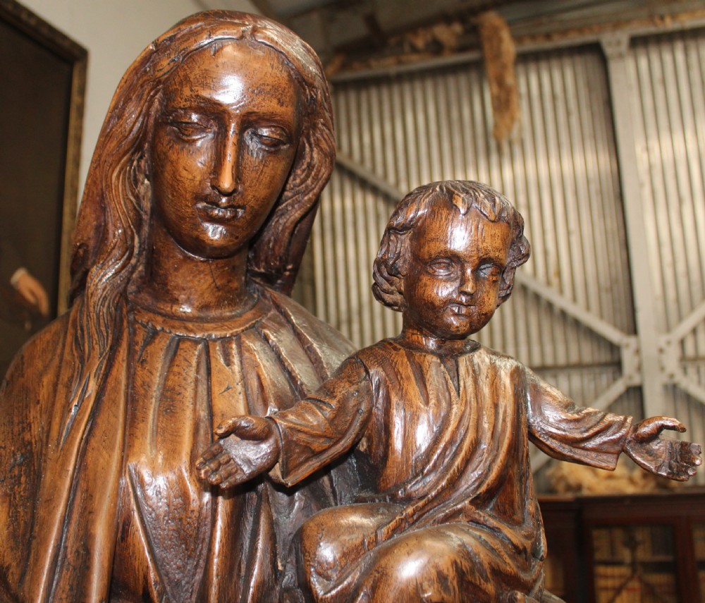 A dark hand-carved wooden statue depicting the Virgin Mary standing and holding the infant Christ in draped robes. View III