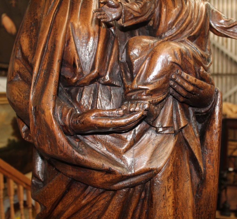 A dark hand-carved wooden statue depicting the Virgin Mary standing and holding the infant Christ in draped robes. View IV