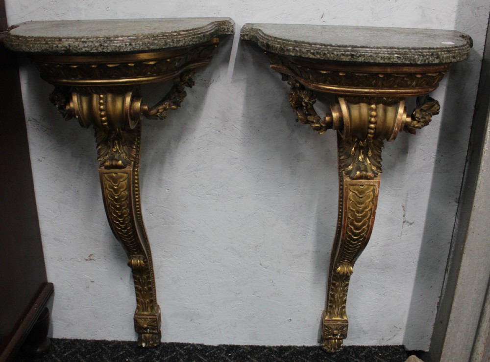 A pair of wall-mounted giltwood console tables with single carved scroll bracket legs and shaped marble tops.
