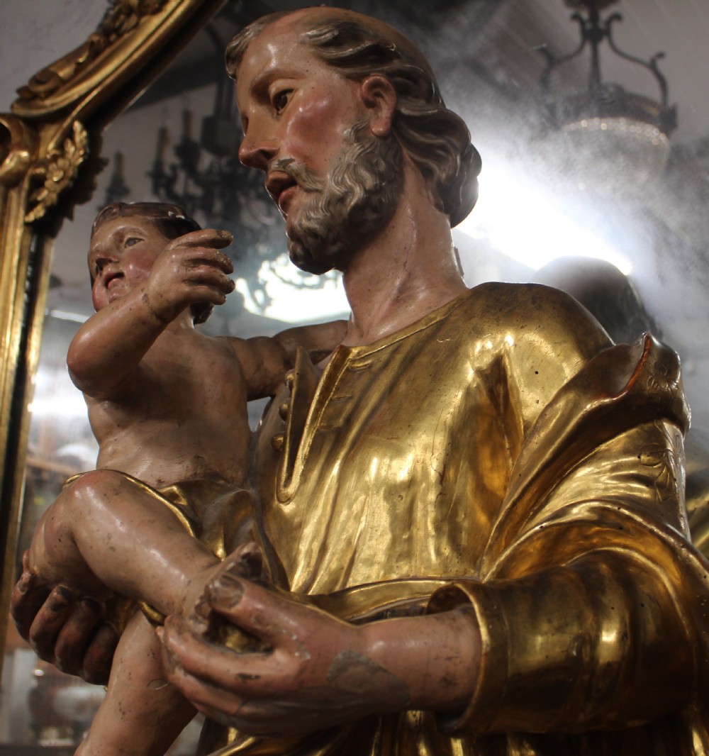 A carved wooden statue depicting Saint Joseph holding the infant Jesus, finished with gold leaf gilding and painted features atop a cherub base. View VI
