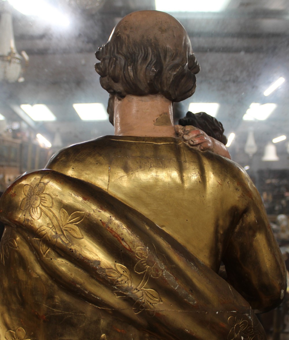 A carved wooden statue depicting Saint Joseph holding the infant Jesus, finished with gold leaf gilding and painted features atop a cherub base. View IX