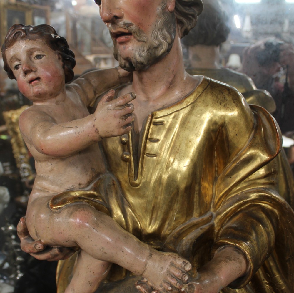 A carved wooden statue depicting Saint Joseph holding the infant Jesus, finished with gold leaf gilding and painted features atop a cherub base. View XIV