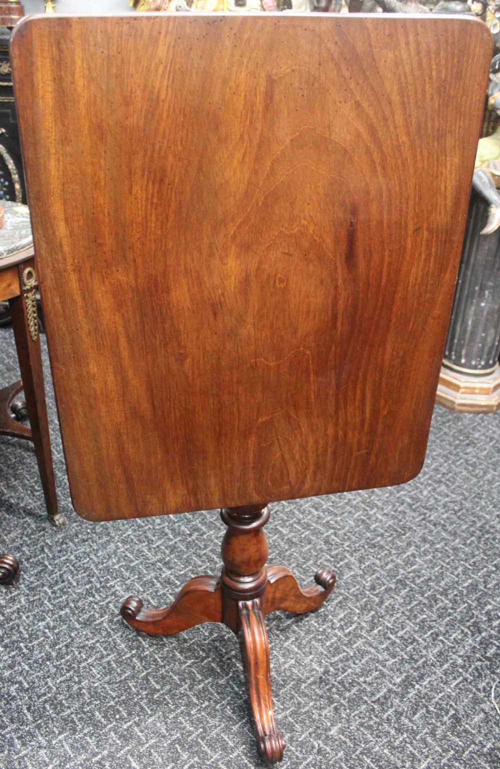 An antique mahogany tilt-top wine table shown with its top tilted upright on a turned pedestal stem and tripod scrolled legs.