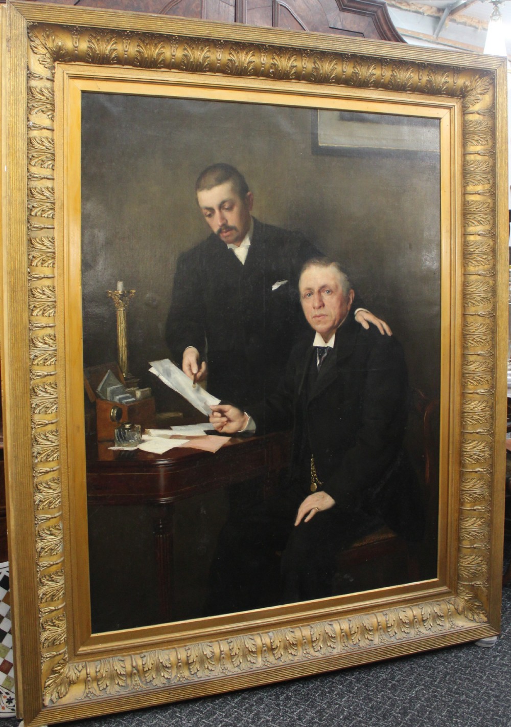 A large antique oil painting depicting two gentlemen in dark suits examining documents near a desk, set in an ornate gilded frame.