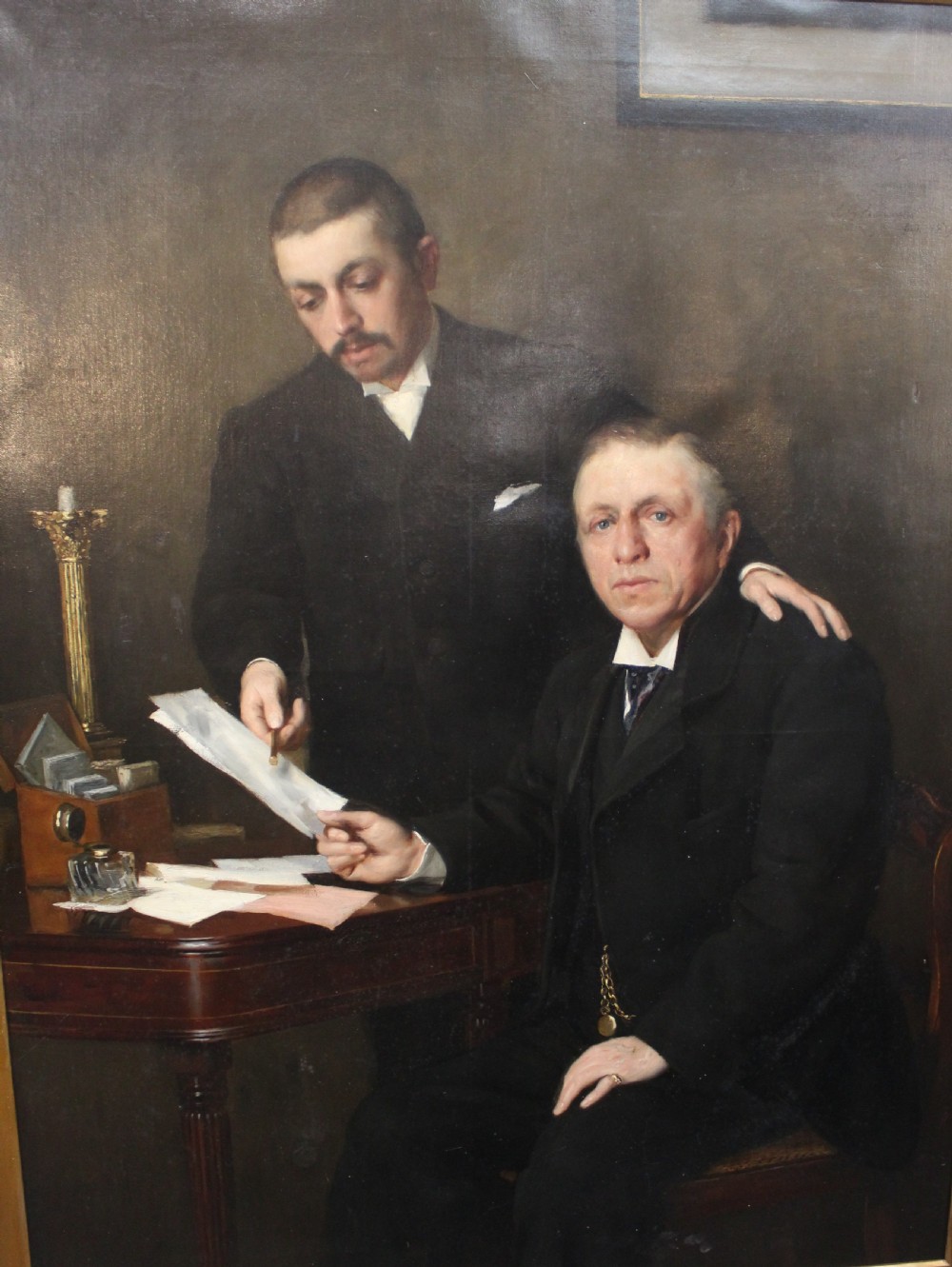 A large antique oil painting depicting two gentlemen in dark suits examining documents near a desk, set in an ornate gilded frame. View V