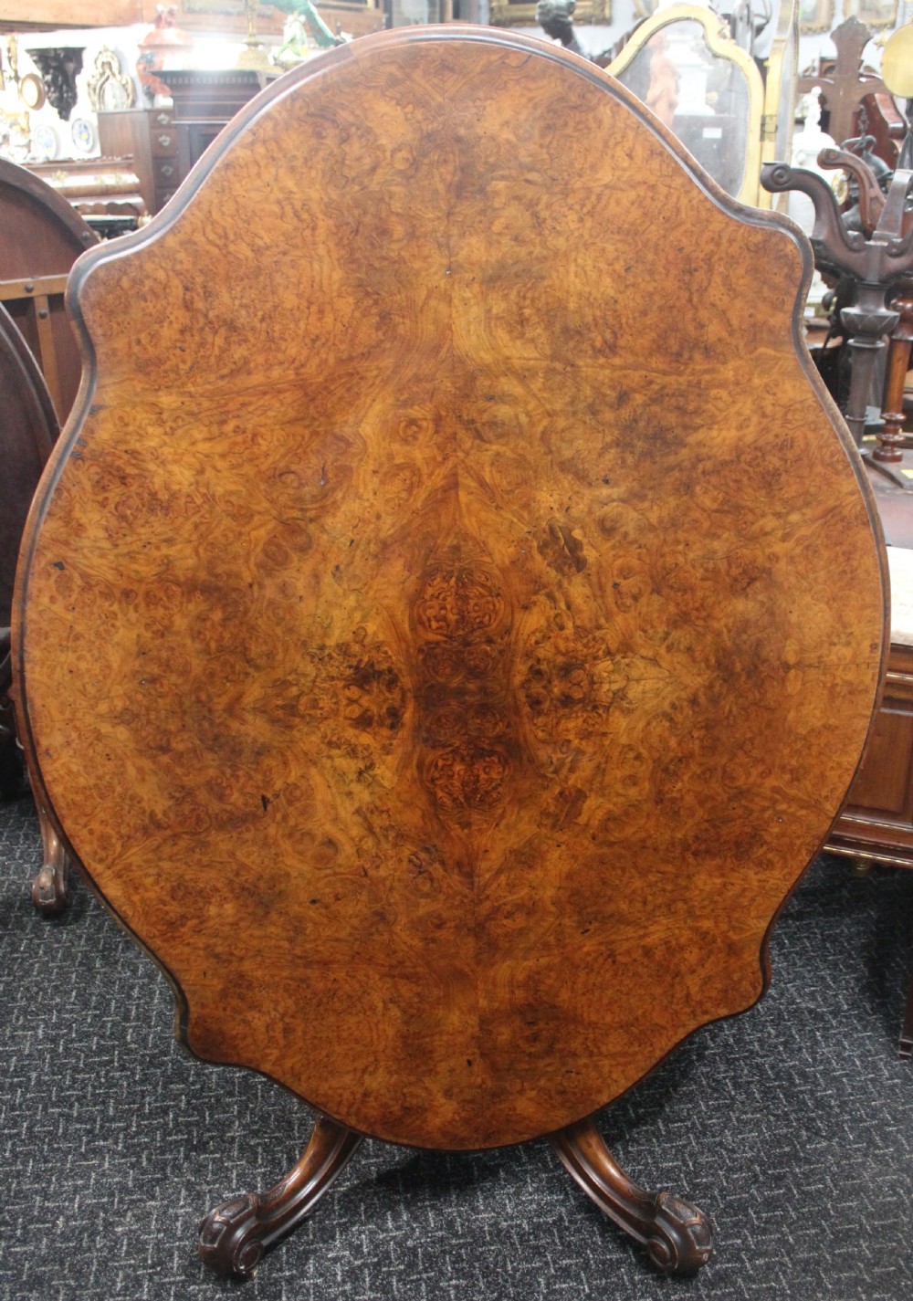 A Victorian burr walnut tilt-top loo table featuring a book-matched serpentine top set on a carved pedestal base with four scrolled legs.