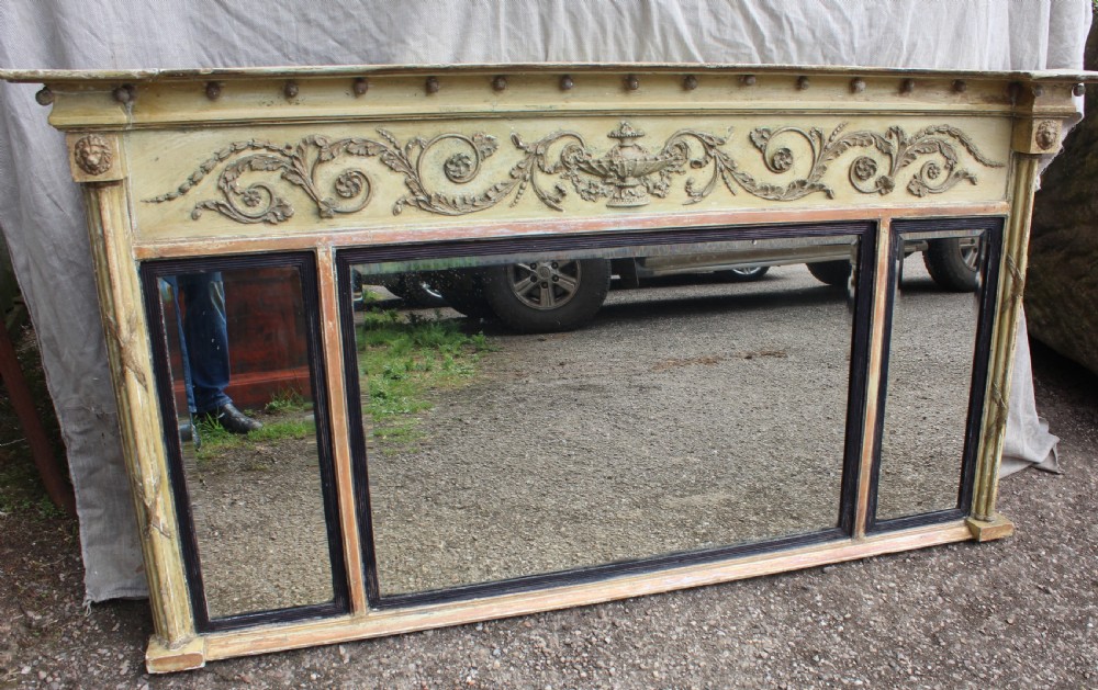 A wide horizontal antique overmantel mirror featuring a distressed cream painted finish, carved neoclassical urn motifs, and three distinct mirror panels.