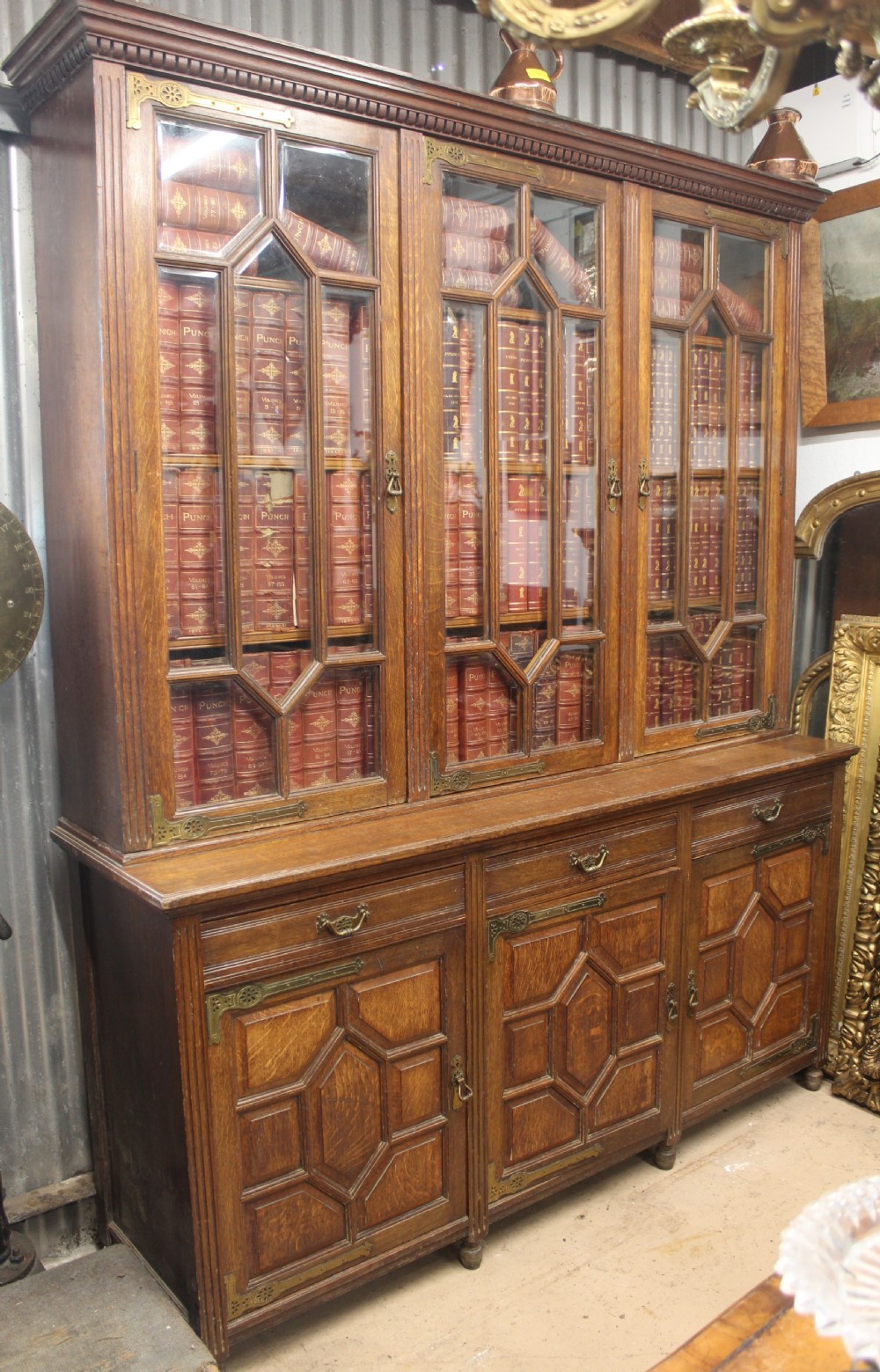 A large English oak glazed bookcase featuring three upper glass doors with geometric mullions above three drawers and three geometric panelled cupboard doors with brass mounts.