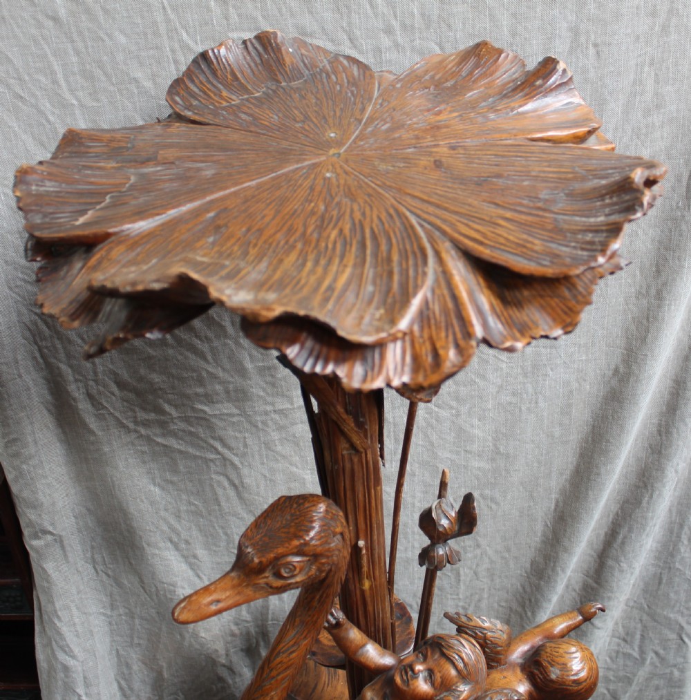 An intricately carved brown wooden pedestal stand featuring putti riding an ostrich surrounded by foliage, topped with a lily pad shaped platform. View III