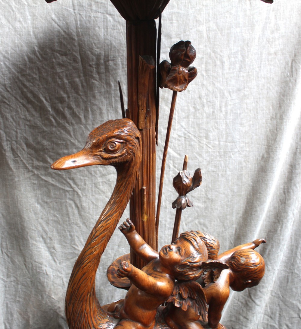 An intricately carved brown wooden pedestal stand featuring putti riding an ostrich surrounded by foliage, topped with a lily pad shaped platform. View IV