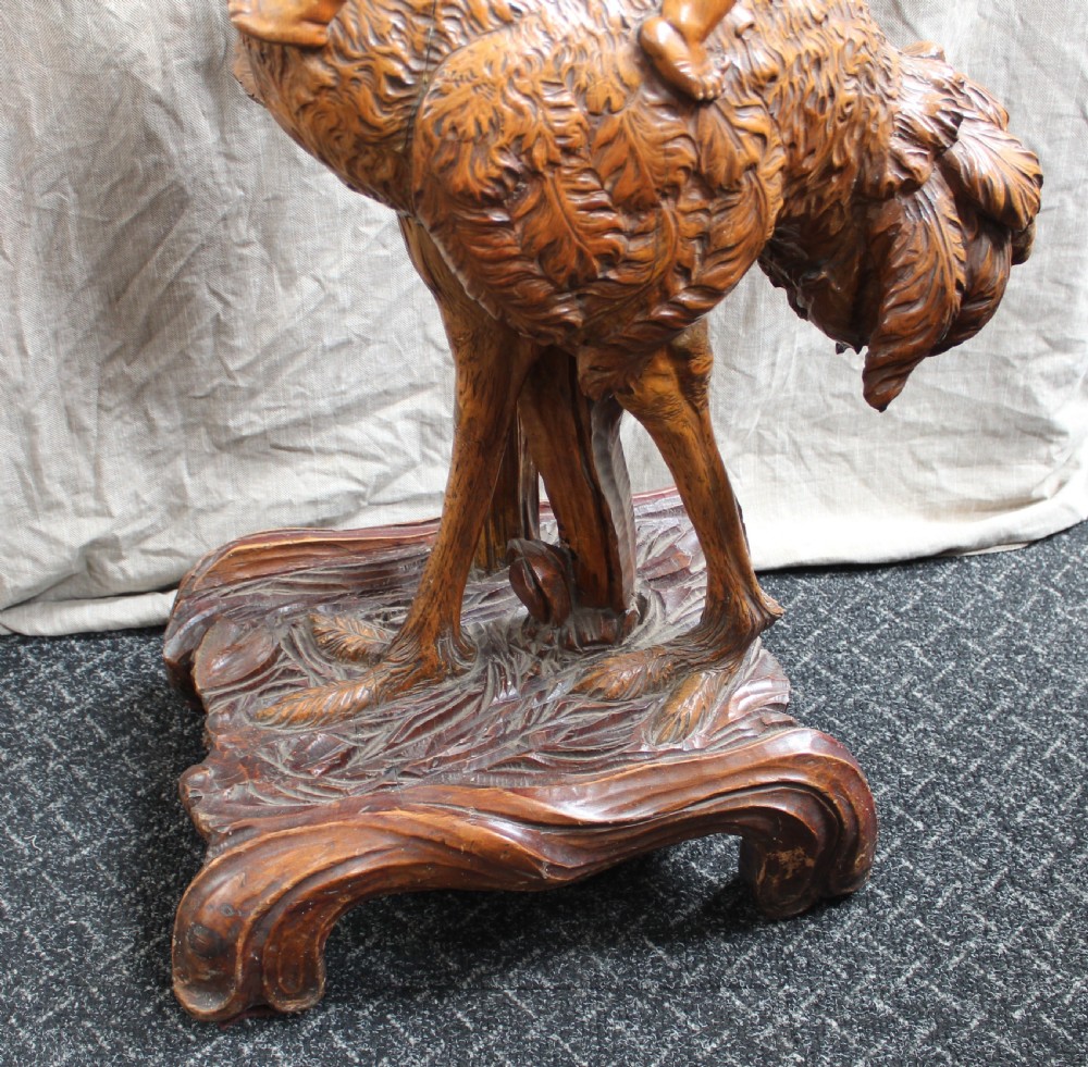 An intricately carved brown wooden pedestal stand featuring putti riding an ostrich surrounded by foliage, topped with a lily pad shaped platform. View V