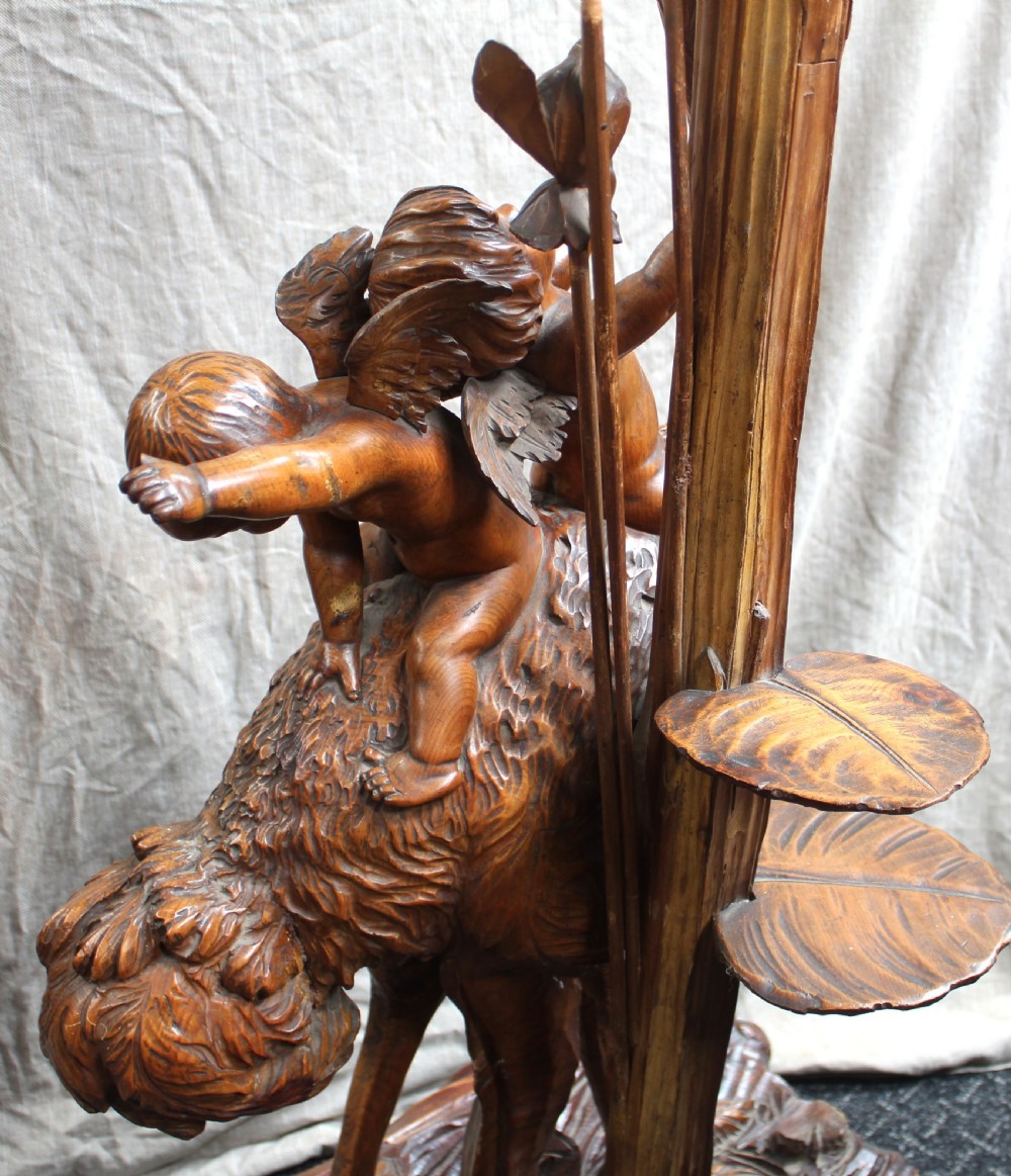 An intricately carved brown wooden pedestal stand featuring putti riding an ostrich surrounded by foliage, topped with a lily pad shaped platform. View VI