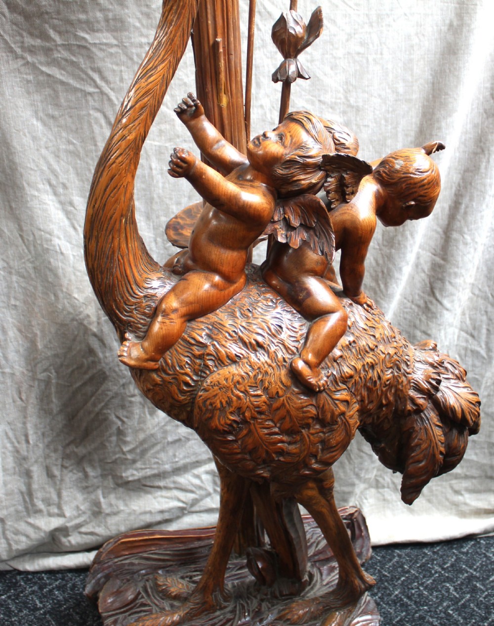 An intricately carved brown wooden pedestal stand featuring putti riding an ostrich surrounded by foliage, topped with a lily pad shaped platform. View VII