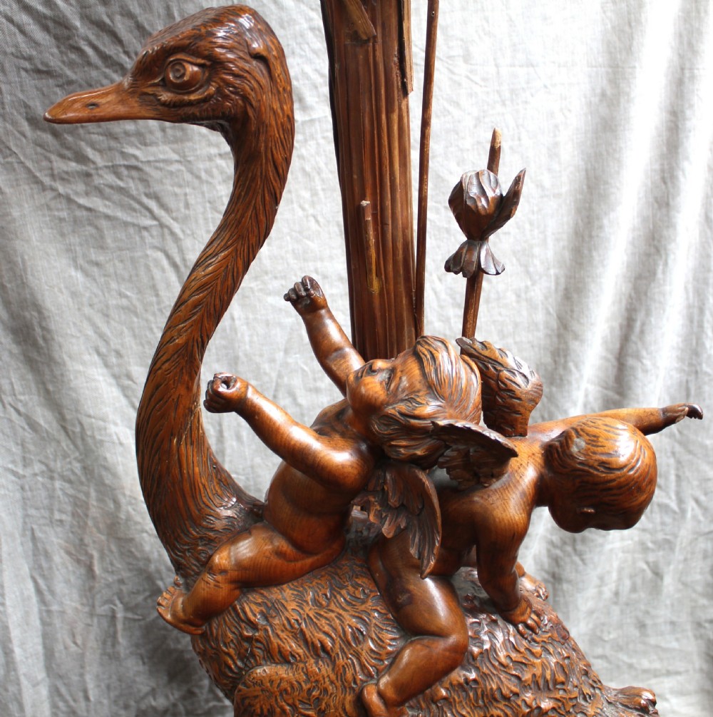An intricately carved brown wooden pedestal stand featuring putti riding an ostrich surrounded by foliage, topped with a lily pad shaped platform. View IX