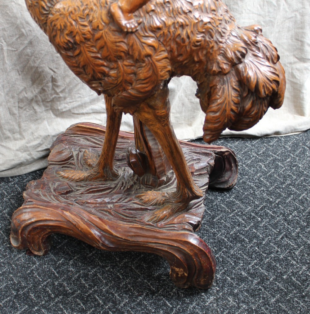 An intricately carved brown wooden pedestal stand featuring putti riding an ostrich surrounded by foliage, topped with a lily pad shaped platform. View X