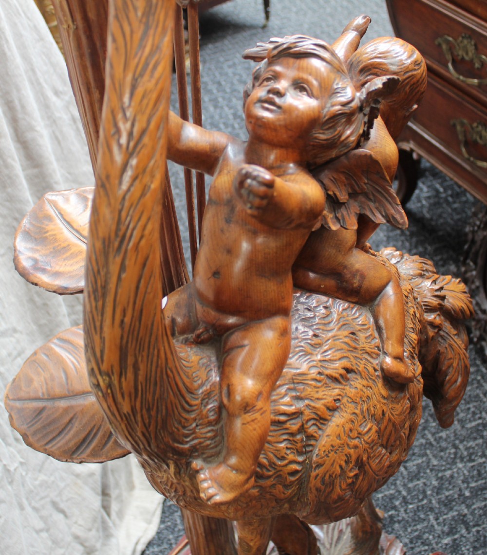 An intricately carved brown wooden pedestal stand featuring putti riding an ostrich surrounded by foliage, topped with a lily pad shaped platform. View XI