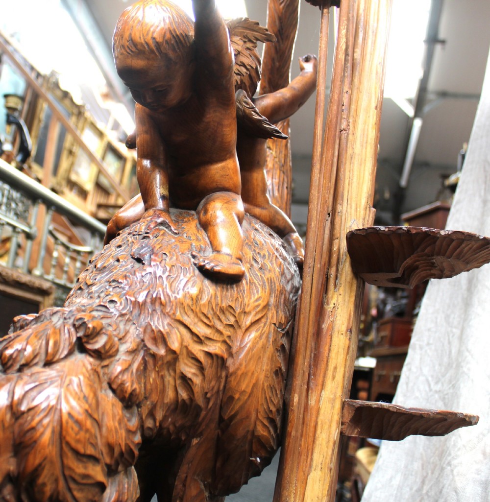 An intricately carved brown wooden pedestal stand featuring putti riding an ostrich surrounded by foliage, topped with a lily pad shaped platform. View XII