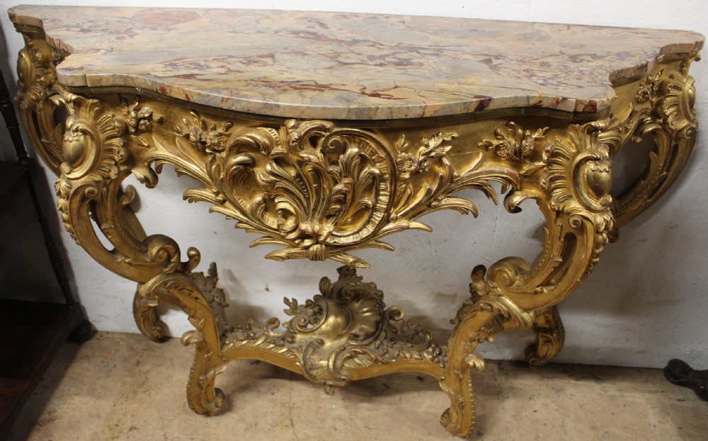 A Rococo style giltwood console table featuring ornate acanthus leaf carving, C-scroll legs, a central shell motif stretcher, and a serpentine variegated marble top.