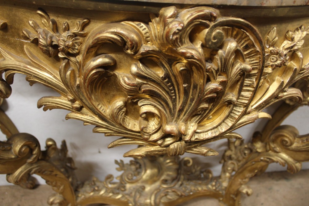 A Rococo style giltwood console table featuring ornate acanthus leaf carving, C-scroll legs, a central shell motif stretcher, and a serpentine variegated marble top. View V