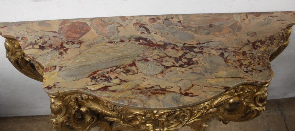 A Rococo style giltwood console table featuring ornate acanthus leaf carving, C-scroll legs, a central shell motif stretcher, and a serpentine variegated marble top. View VII