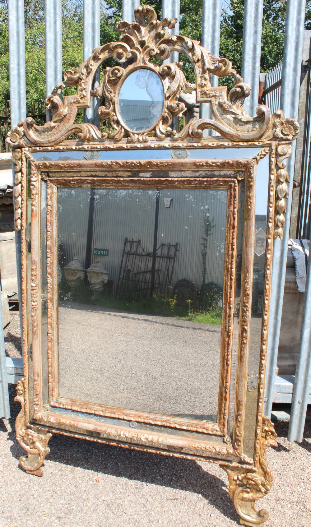 An antique Italian gilded wood mirror featuring a carved ornate crest with a small oval mirror, framed by mirrored glass borders and scroll feet.