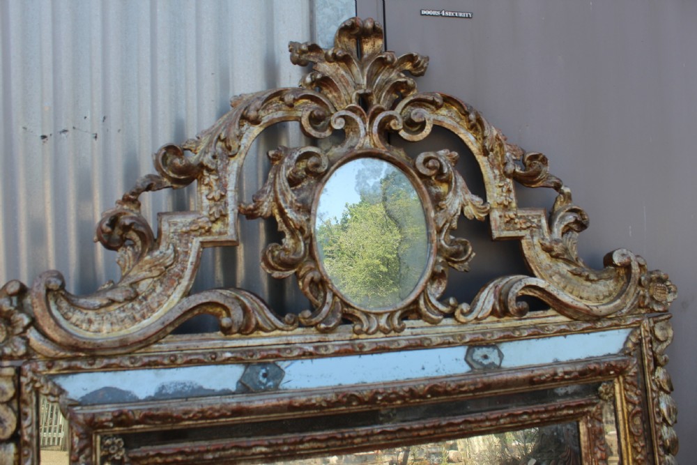 An antique Italian gilded wood mirror featuring a carved ornate crest with a small oval mirror, framed by mirrored glass borders and scroll feet. View III