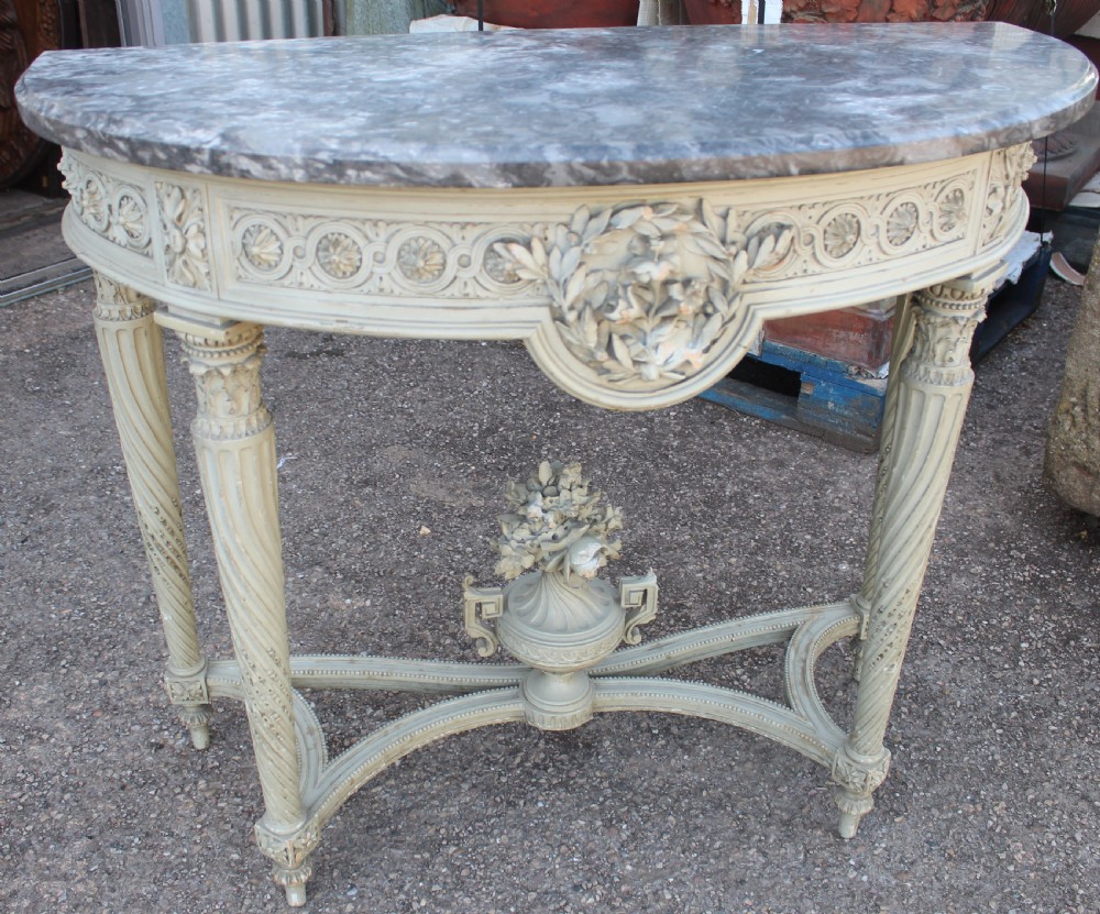 A Neoclassical French Louis XVI style demilune console table featuring a grey marble top, painted off-white carved apron, spiralled fluted legs, and a stretcher centred with a carved urn.