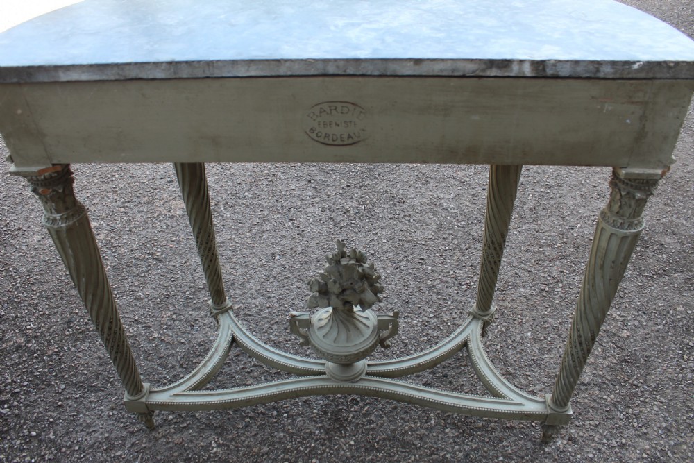 A Neoclassical French Louis XVI style demilune console table featuring a grey marble top, painted off-white carved apron, spiralled fluted legs, and a stretcher centred with a carved urn. View VI