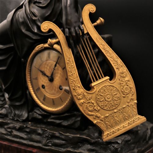 fig 2. A French Empire mantel clock featuring a dark patinated bronze classical female figure holding a lyre, positioned beside a gold-coloured dial on a red marble base with gilt metal mounts.