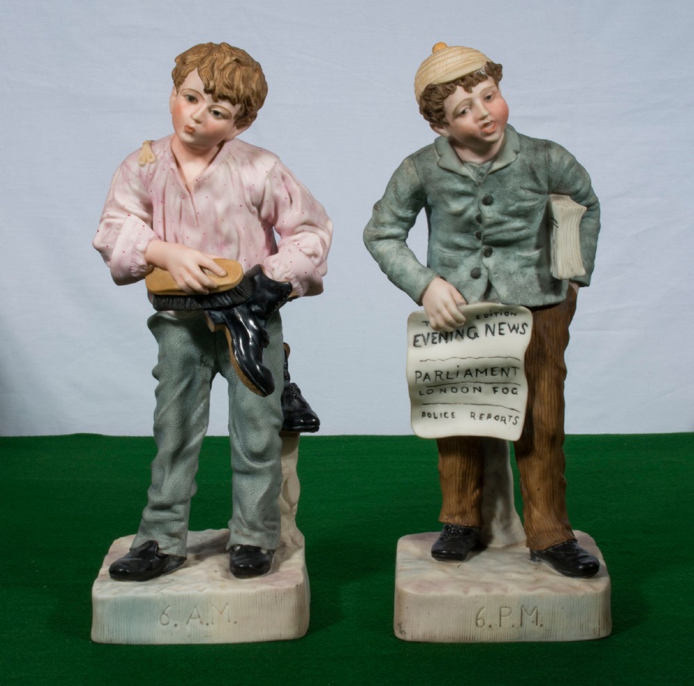 A pair of antique painted bisque porcelain figurines depicting two young boys, inscribed 6 A.M. polishing a shoe and 6 P.M. holding a newspaper.