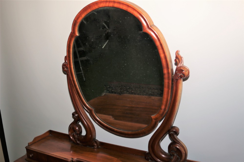 A Victorian flame mahogany Duchess dressing table featuring an oval swivel mirror, upper trinket drawers, and carved scroll legs. View VI
