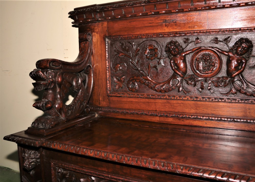 A dark carved walnut hall seat featuring high-relief carvings of mythical figures and floral scrollwork on the backrest and front storage panel. View VIII