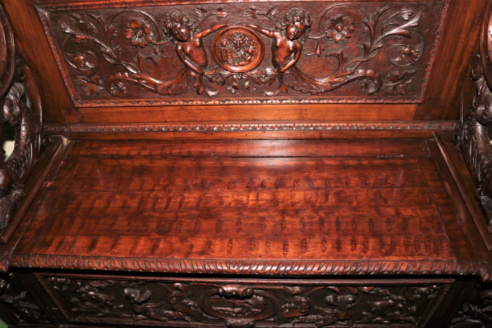 A dark carved walnut hall seat featuring high-relief carvings of mythical figures and floral scrollwork on the backrest and front storage panel. View XIV