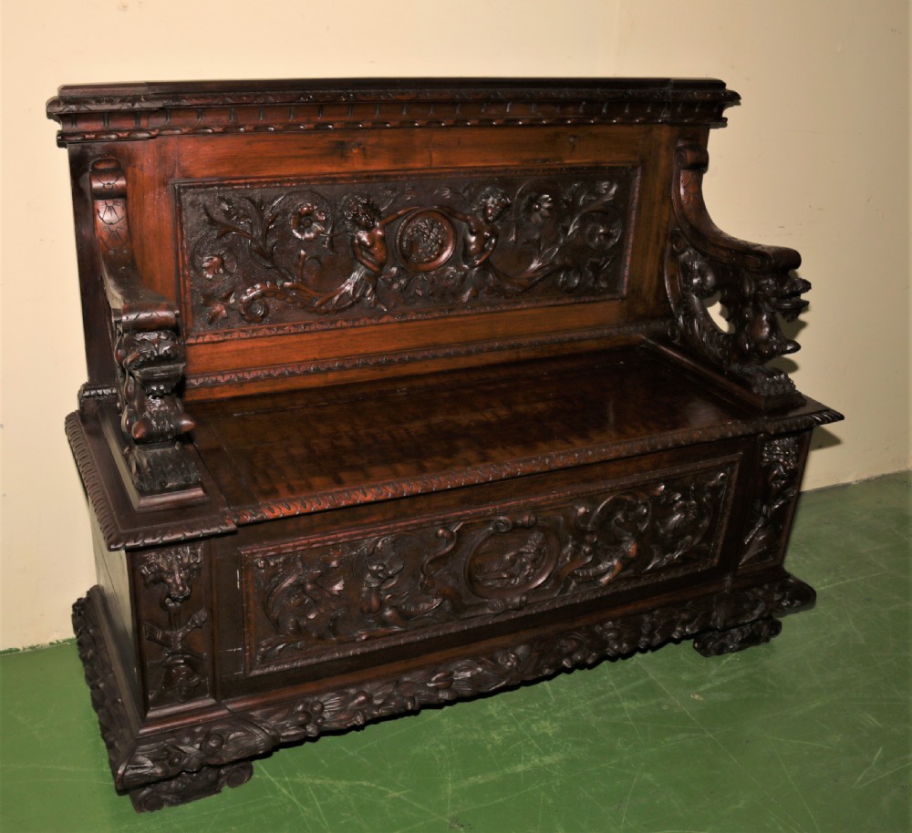 A dark carved walnut hall seat featuring high-relief carvings of mythical figures and floral scrollwork on the backrest and front storage panel. View XVI